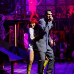 A performer in a red wig and yellow boots strikes a dynamic pose alongside a singer in a suit on a colorful stage set.