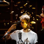 A man with disheveled hair pointing a gun to his head on stage with golden confetti floating around and a puppet character to his side.