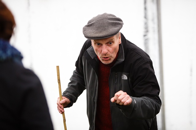 A man in a flat cap and jacket gestures emphatically while holding a stick and speaking to someone off-camera.