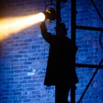 A silhouette of a person adjusting a spotlight on a scaffold against a brick wall.