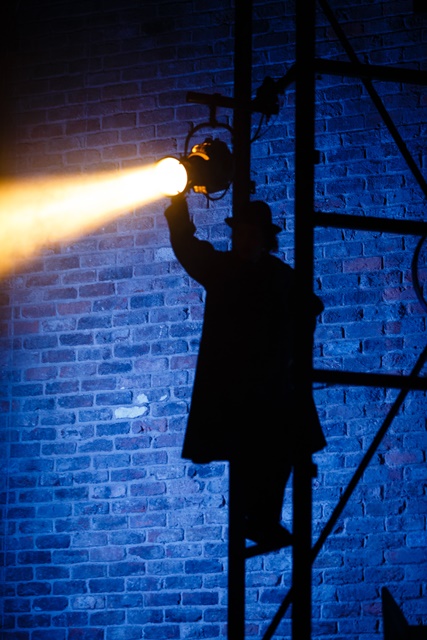 A silhouette of a person adjusting a spotlight on a scaffold against a brick wall.