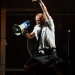 Man in a kilt jumping with a megaphone on stage.