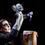 A puppeteer performs with a puppet dressed as a scientist holding a book, illuminated under a spotlight.