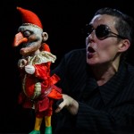 A performer in black attire and sunglasses vocalizing beside a puppet clown in a red costume.