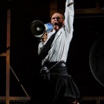 A man in a kilt and white shirt dramatically speaking into a megaphone on stage.