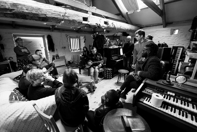 Group of people gathered in a cozy, rustic music studio with instruments visible.