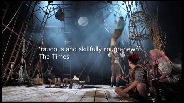A group of actors on a rustic set performing in a theatrical production, with a review quote from the times displayed on the screen.
