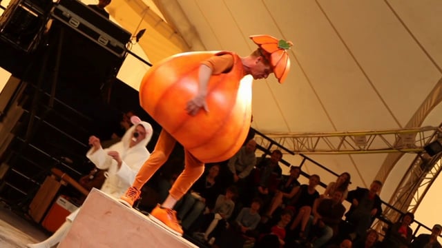 Performer in an orange pumpkin costume on stage at an event.
