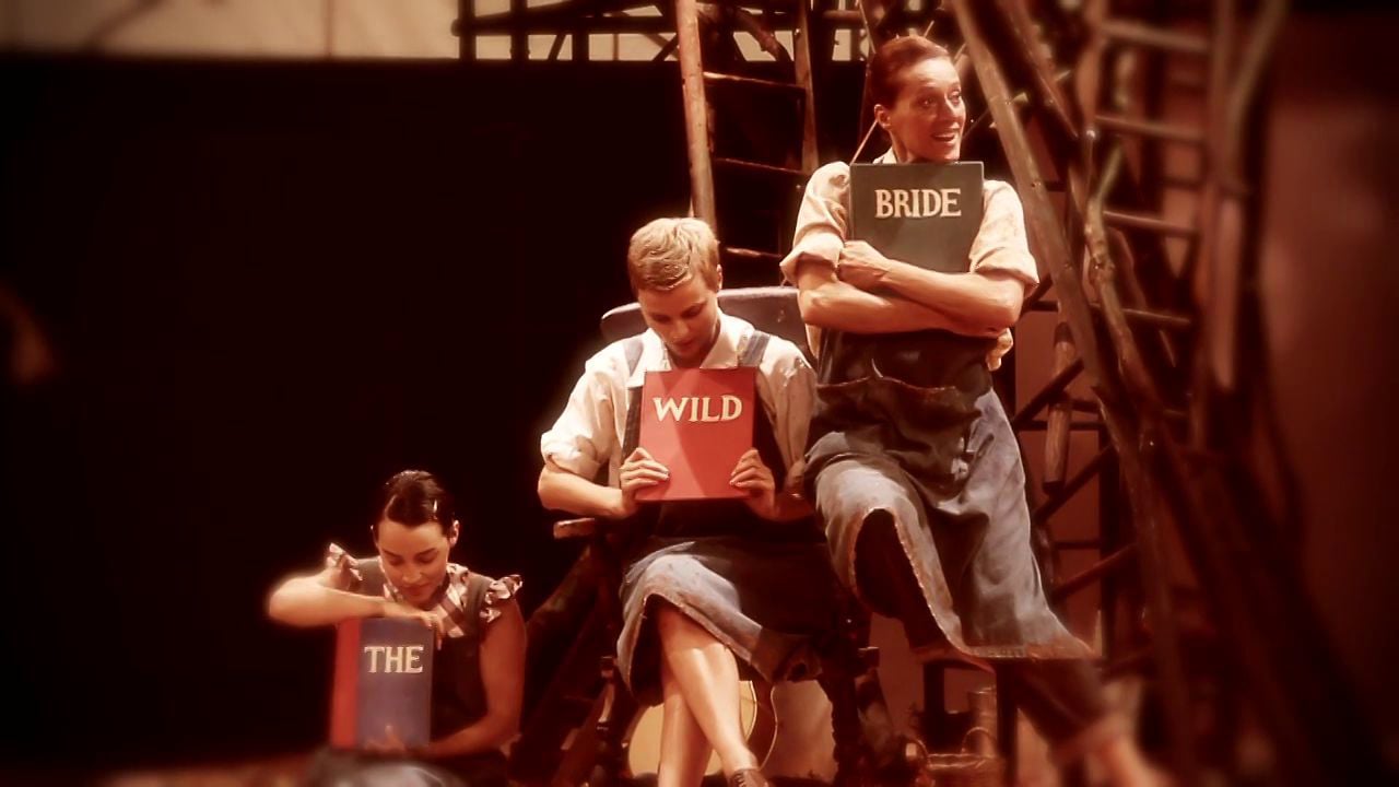Three performers on stage with props, one holding a sign that reads 'wild', another 'the', and the one in the center 'bride'.