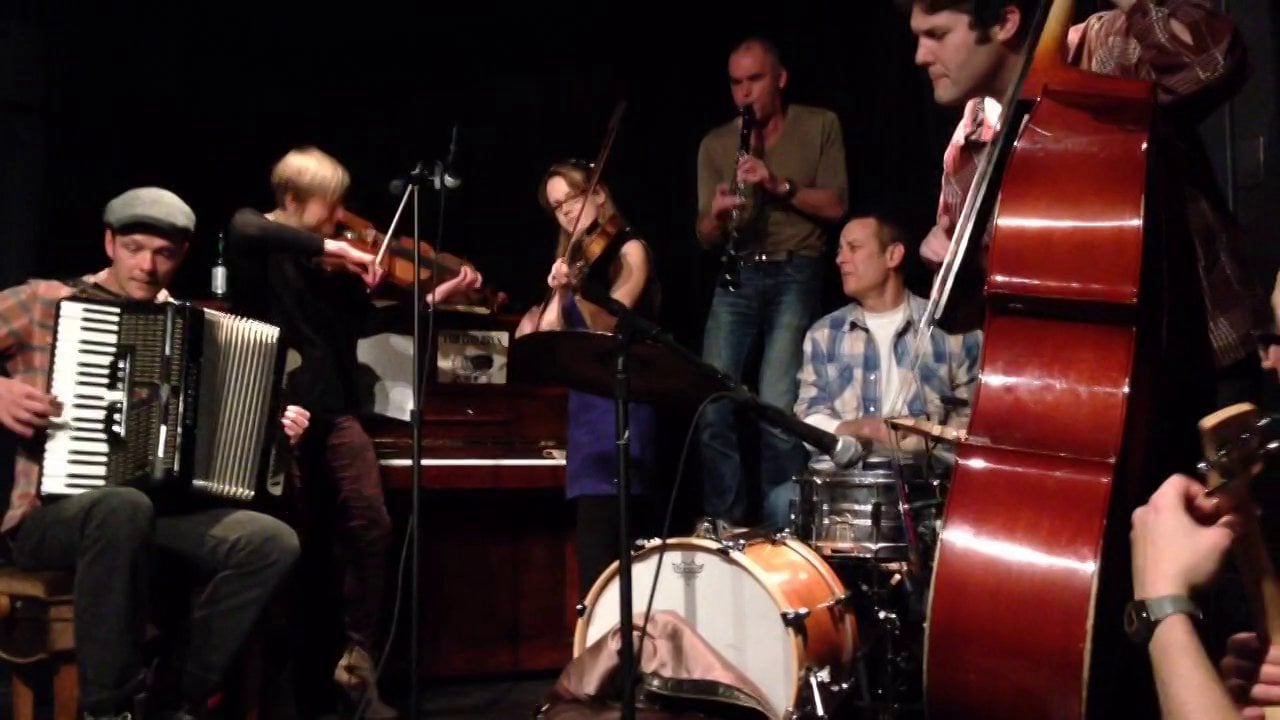 A group of musicians performing, featuring instruments such as an accordion, violin, double bass, and drums.