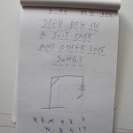 Notepad with handwritten song title 'dead dog in a suitcase' and other text, with a simple drawing and stick figures.
