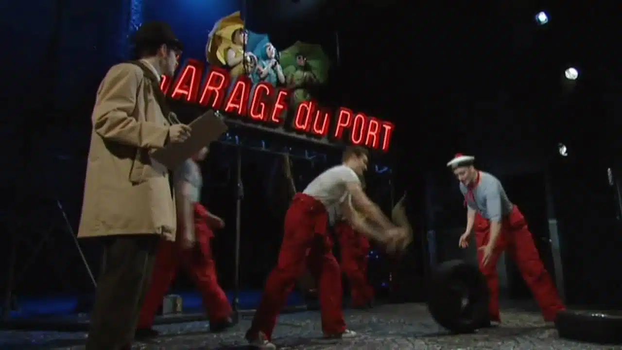 Three performers on a stage with one dressed in a trench coat reading a newspaper, and two others in red overalls playfully interacting with a tire under a neon sign that reads 'garage du port.