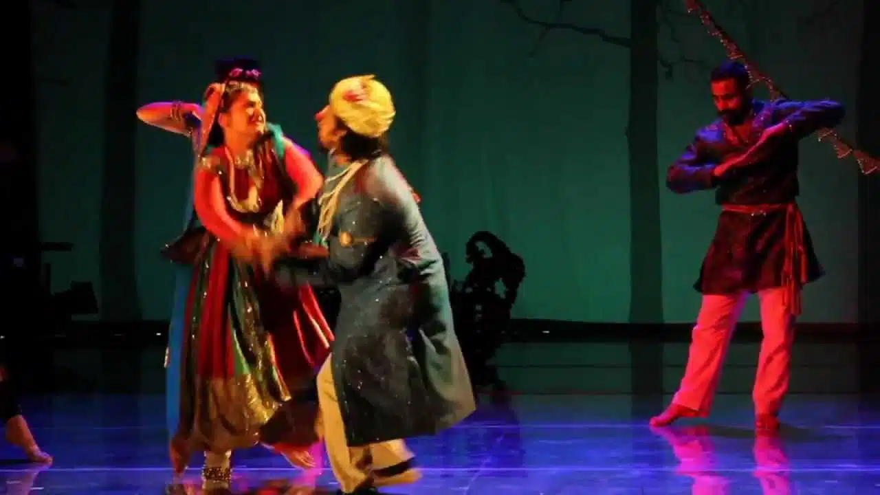 Two performers in traditional attire dancing on stage with another person in the background.