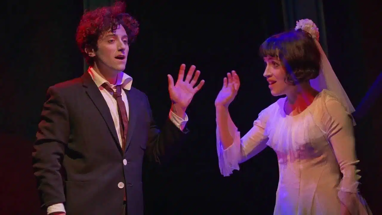 Two actors performing on stage, with the man looking surprised and the woman gesturing animatedly.