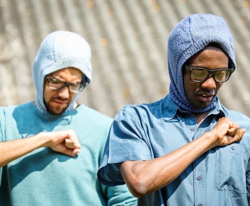 Featured image for 'Tristan and Yseult - Rehearsal Images'. The image contains two individuals from the chest up dressed in blue shirts and blue wool hoods. They are standing outside in daylight and holding their fists over their hearts. They are looking down with a solemn look on their faces and are both wearing black rimmed glasses