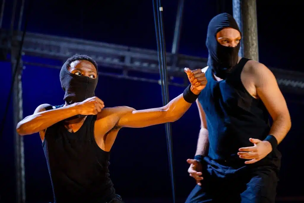 Two characters - the brutes - stand in black vests with balaclava masks over their faces. The brute on the left has his fists raised