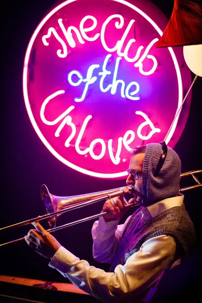 A love spotter holding an instrument with a neon sign reading 'The Club of the Unloved' directly behind him