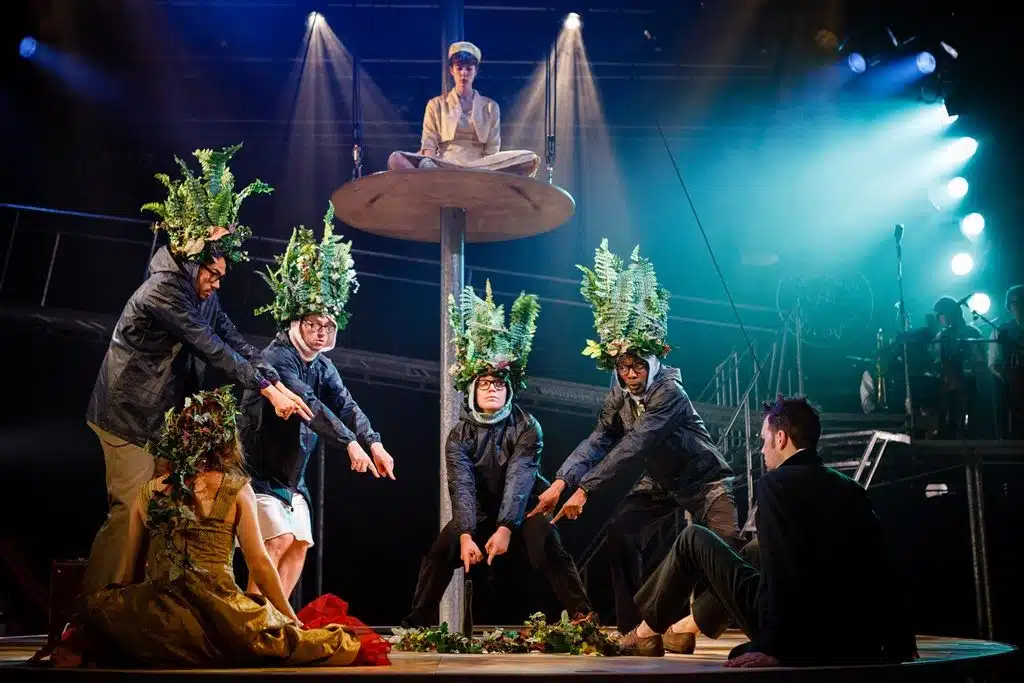 Tristand and Yseult sit with their back to the camera as they are faced with four love spotters stood in front of them, each with foliage head coverings. The four are pointing towards a pile of foliage on the floor. Whitehands sits on a platform above this, out of view