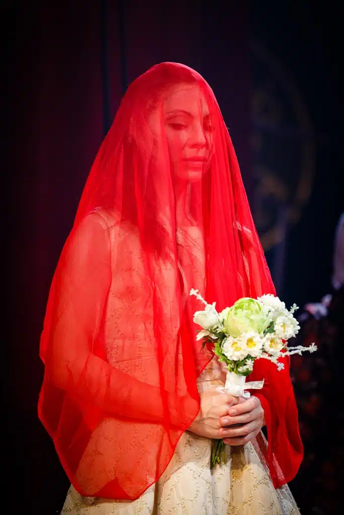 Yseult stands in a gold dress, clasping a small bouquet of white flowers, with a red veil over her head