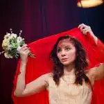 Yseult stands in a gold gown holding a red sheet above her head, and a small bouquet of white blowers in her left hand