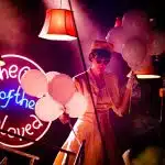 Whitehands stands in ominous red righting with a focused expression, holding bunches of balloons in each hand. There are multiple lampshades in the shot which provide low light, and there is a neon sign in the background with reads 'The Club of the Unloved'