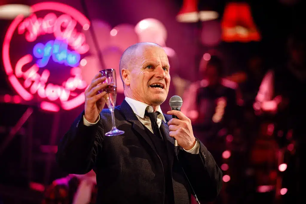 King Mark stands in the centre foreground in a black suit, speaking into a microphone with a glass of champagne in his other hand. To his upper left is a sign which reads 'Club of the Unloved'