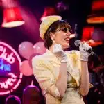 Whitehands stands in the foreground in her 1950s style yellow dress and sunglass. She is singing into a microphone in one hand, with a cigarette in her other hand. To her left is a neon sign which reads 'The Club of the Unloved'.