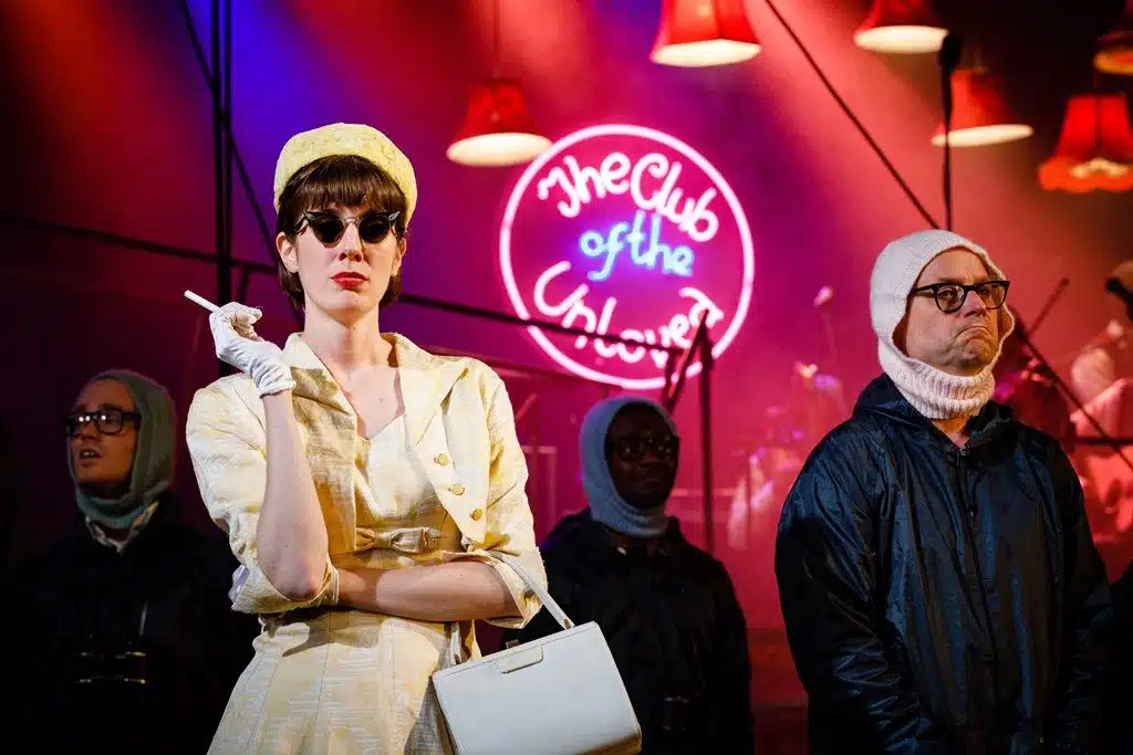 Whitehands stands in the foreground in her 1950s style yellow dress and sunglass, with her arms crosses and a cigarette in one hand near her mouth. Three of the Love Spotters stand behind her, and abve them is a neon sign which reads 'The Club of the Unloved'.