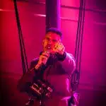 Frocin sings with a microphone in one hand, the other pointing towards the camera. He is wearing a harness and has a polaroid camera around his neck. The scene lighting is very pink