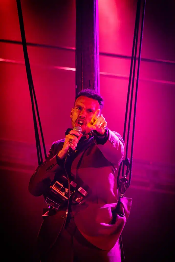Frocin sings with a microphone in one hand, the other pointing towards the camera. He is wearing a harness and has a polaroid camera around his neck. The scene lighting is very pink