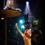 Yseult dances on stage with her arms in the air. Two people stand in the background dressed as foliage, with one playing a violin. Whitehands sits on a platform above Yseult, mostly out of view, pointing in the direction of the camera