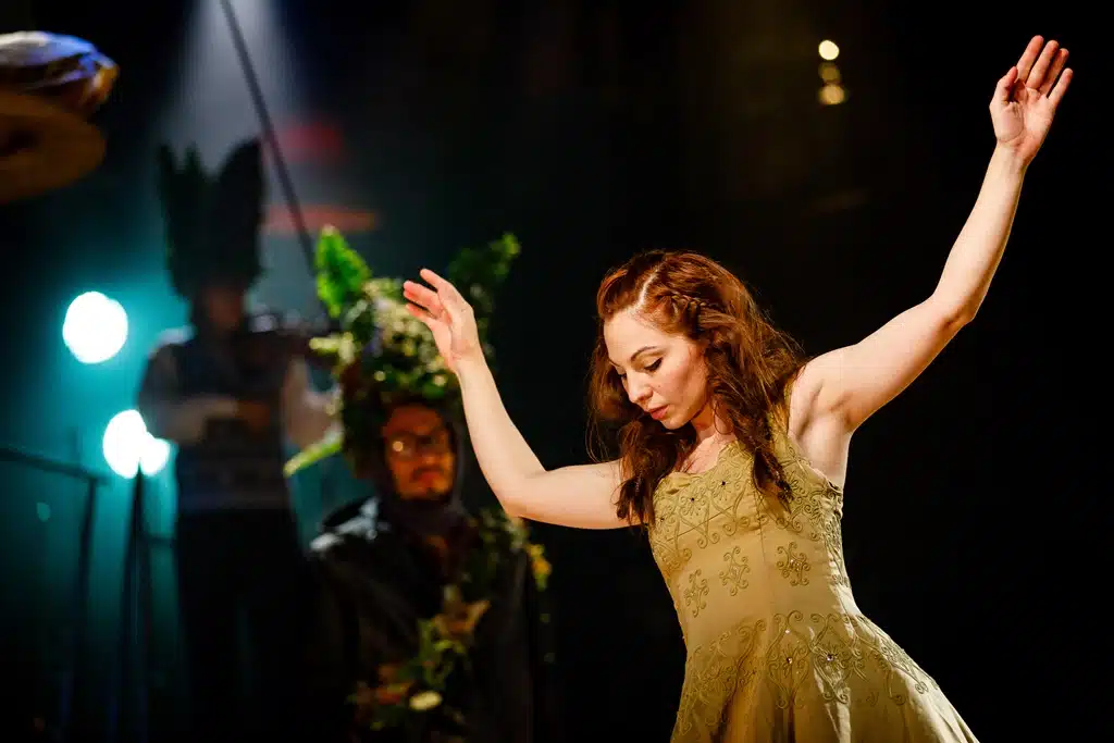 Featured image from 'Production images of Tristan and Yseult'. Yseult dances on stage in a green dress, with a person dressed as foliage in the background