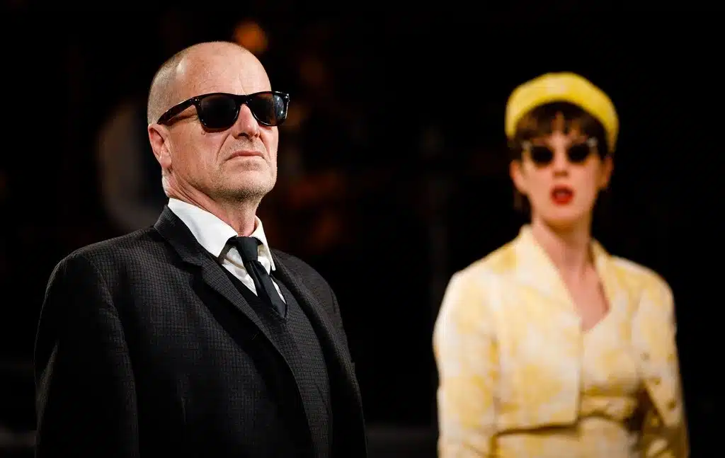 King Mark on the left stands on the left with an unreadable expression, in a black suit and sunglasses. Whitehands stands on the right slightly behind him. She wears a yellow 1950s style dress and hat, with sunglasses and red lipstick