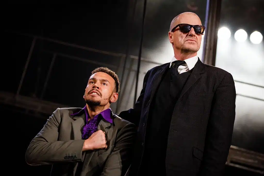 Frocin & King Mark. Frocin, on the left, stands with his fist held to his chest, wearing a grey suit and purple shirt. King Mark stands tall beside him, looking away, wearing a black suit and sunglasses