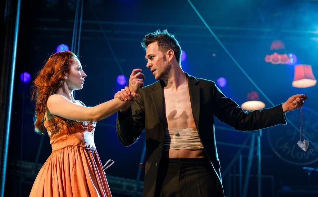 Dominic March as Tristan & Hannah Vassallo as Yseult. Yseult has her hand outstretched to Tristan's face, who has a grasp of her wrist. Tristan looks dishevelled, with wounds on his face, and his open blazer reveals bandages around his torso. In his other hand he holds a heart necklace away from them