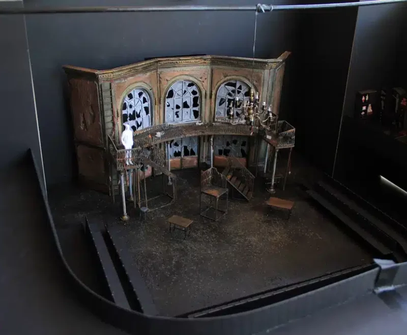 Photograph for Kneehigh's The Tin Drum. Image features a model box which has a building wall with broken windows. There is also a chandelier and staircase with white figure, below is a chair and two small tables.