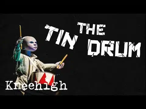 A thumbnail image for a trailer for Kneehigh's Tin Drum. The image is of a small porcelain doll with an angry look on its face. The doll is dressed in a simple white coat and it holding two drum sticks in the air. A small red and white drum is hung around his neck by a strap. There is some bold white text with slight cracks in the font which reads 'THE TIN DRUM'. Both the text and the doll are backed by a plain black background.