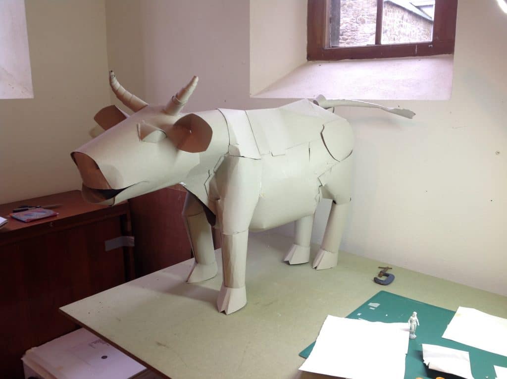 A photo of a prop bull made from white paper and cardboard placed on a table. The bull is roughly a foot tall with elaborate details like a tail, hoofs and horns.