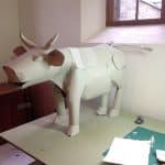 A photo of a prop bull made from white paper and cardboard placed on a table. The bull is roughly a foot tall with elaborate details like a tail, hoofs and horns.
