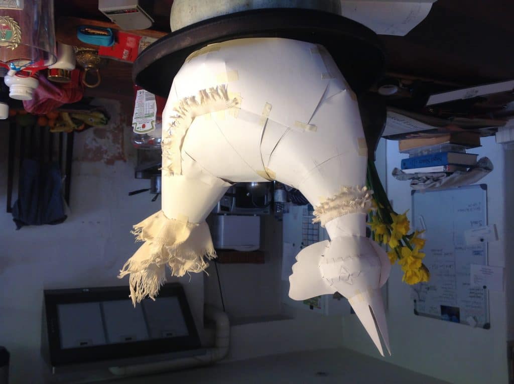 An upside down image of a sculpture of a bird made out of paper in a cluttered room. Books, utilities and general household items can be seen in the background.