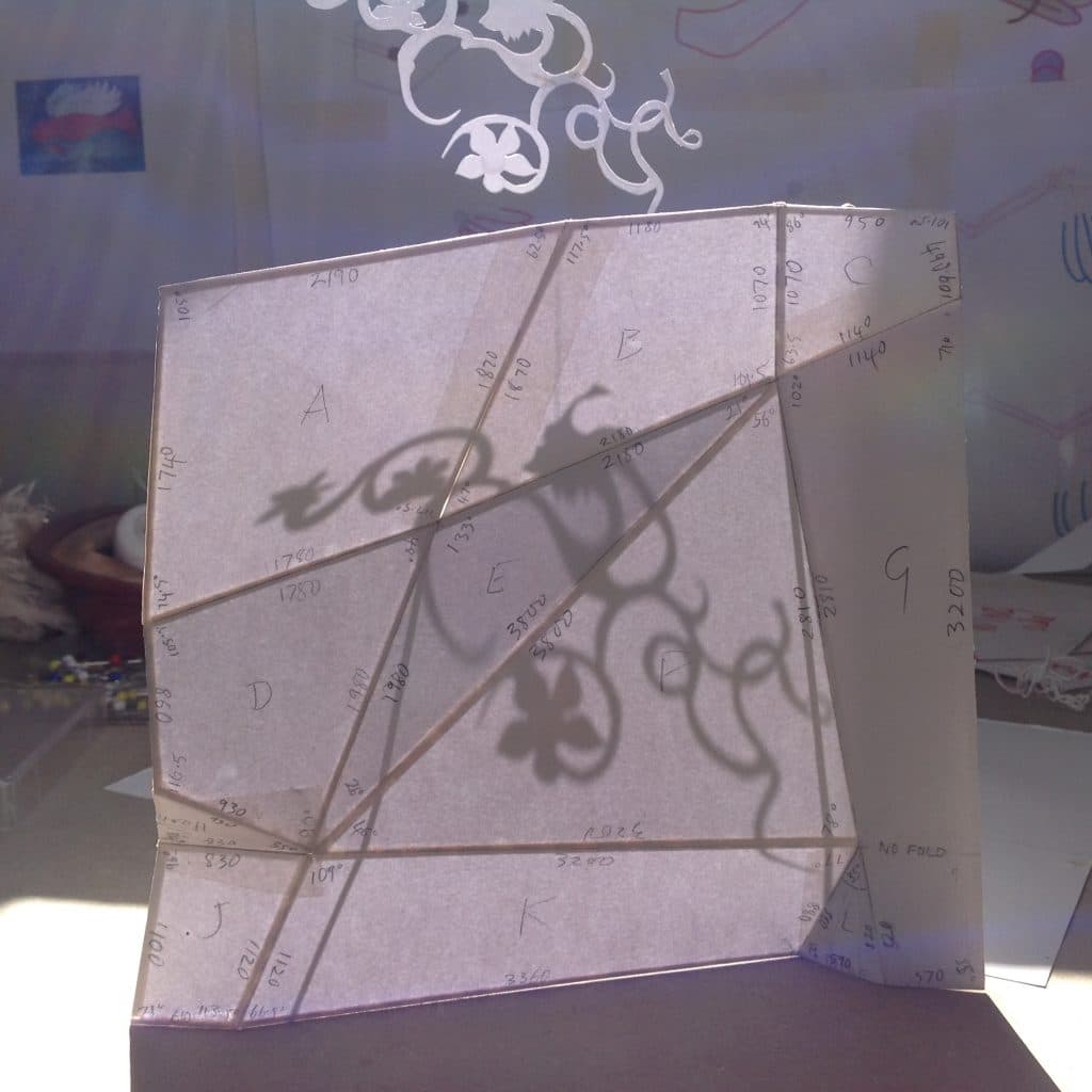 A small prop or design made of white papers and small wooden sticks. There is a floral shadow cast on the prop by a nearby hanging item. A rough white small with small illustrations forms the backdrop.