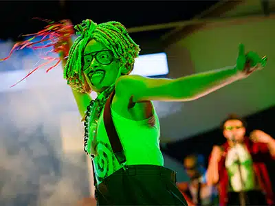 Photograph from Kneehigh's Ubu. Image depicts Katy Owen as Ubu wearing a costume that consists of black trousers, white top with black spiral and red trouser braces. Katy Owen is being lit up by a green light and has handful of red ribbons. The Sweaty Bureacrats band can be seen in the background.