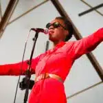 Photograph for Kneehigh's Ubu. Image features Nandi Bhebhe dressed in red and wearing black sunglasses, singing on stage.