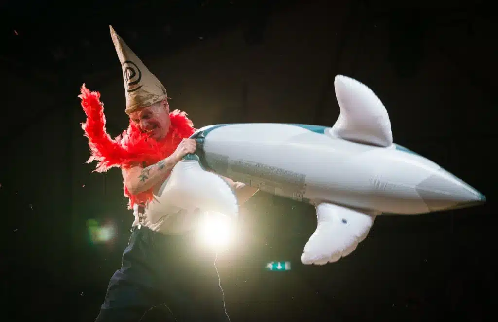 Photograph from Kneehigh's Ubu. The image features Mike Shepherd as Mrs Ubu standing on stage holding an inflatable dolphin amidst a black background. He is dressed in full costume consisting of black trousers, white top, red scarf and a pointy hat with a spiral.