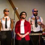 Photograph from Kneehigh's Ubu. The image features 'The Sweaty Bureaucrats band' standing on stage dressed in red, white and black with sunglasses.
