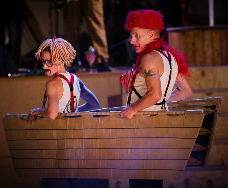 Photograph from Kneehigh's Ubu. Featuring Katy Owen as Ubu and Mike Shepherd as Mrs Ubu. Both are standing on stage in a boat and wearing full costume which is white, black and red.