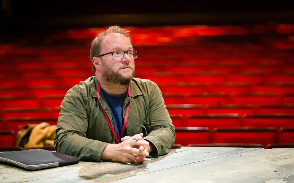 Photograph for Kneehigh's Ubu. Image features Carl Grose sat down in a theatre, rows of red seats can be seen behind him.