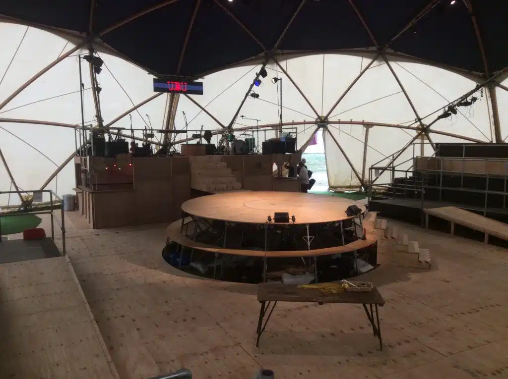 Photograph of Kneehigh's Ubu stage. Circular wooden stage with steps in foreground with musicians stage to the rear with central steps. Musical equipment on musicians stage with striped sign with UBU in bold red hanging above stage. All within geodome tent.