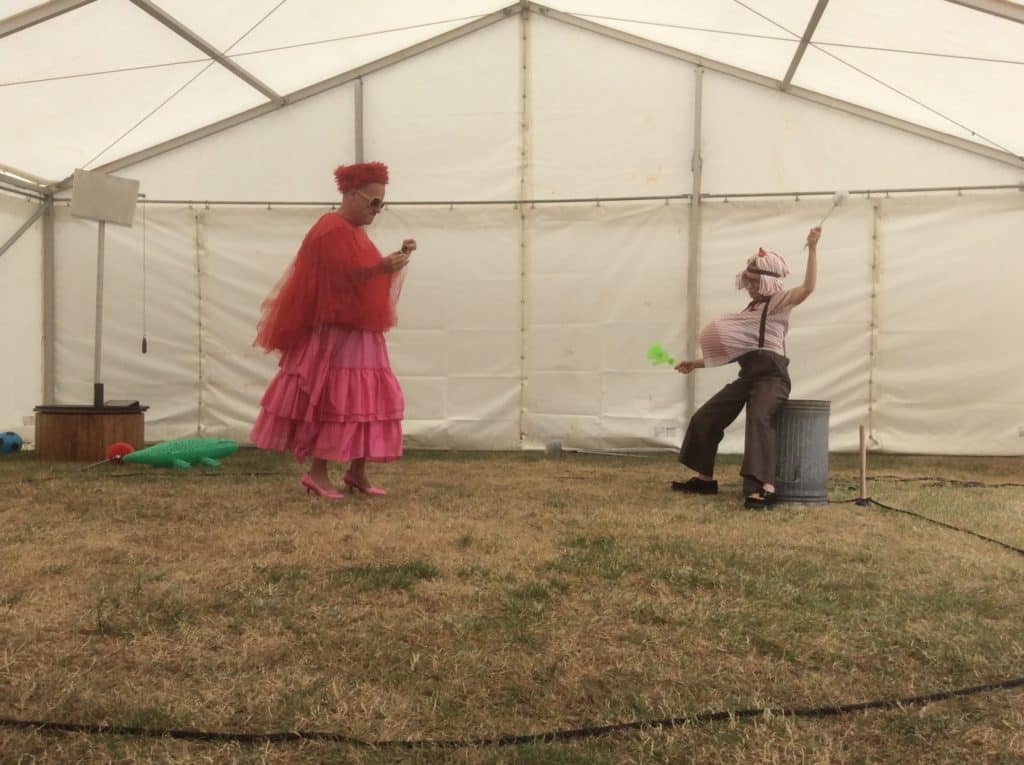 Photograph for Kneehigh's Ubu. Image features a white gazebo with grass on the ground and two performers dressed as Mr and Mrs Ubu. Mrs Ubu is wearing a pink skirt, pink shoes, red flowing top and red hat with sunglasses. Mr Ubu is wearing dark grey trousers, white shirt with trouser brace and object stuffed inside shirt that makes his belly look bigger.Mr Ubu is holding a toilet brush and perched against a metal bin.