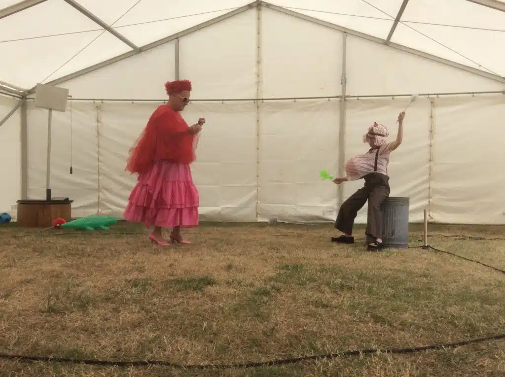 Photograph for Kneehigh's Ubu. Image features a white gazebo with grass on the ground and two performers dressed as Mr and Mrs Ubu. Mrs Ubu is wearing a pink skirt, pink shoes, red flowing top and red hat with sunglasses. Mr Ubu is wearing dark grey trousers, white shirt with trouser brace and object stuffed inside shirt that makes his belly look bigger.Mr Ubu is holding a toilet brush and perched against a metal bin.